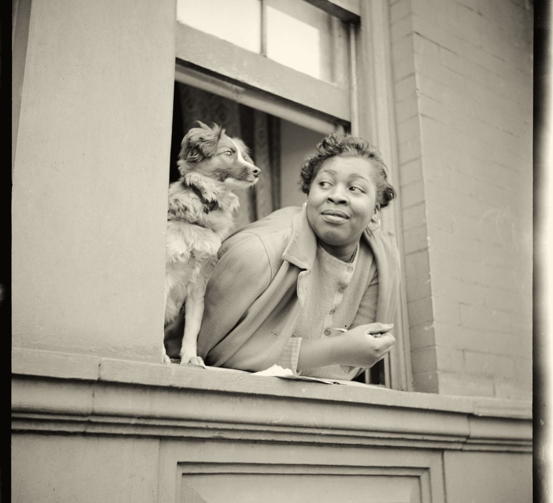 Archival photograph of a woman and her dog leaning out of a rowhouse window